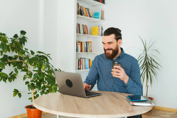 Фото веселого бородатого мужчины, сидящего на рабочем месте и пьющего кофе и смотрящего на ноутбук - Фото, изображение