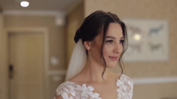 Mooie bruid in witte trouwjurk en sluier thuis blijven in de woonkamer, kijken naar de camera, glimlachen - Video