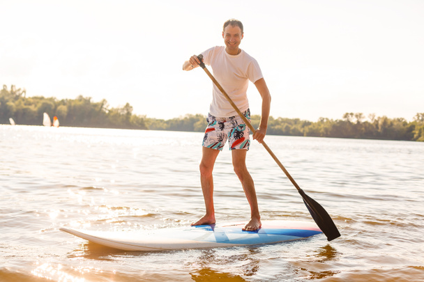 Retrato de un surfista con una tabla SUP en la playa. Joven en el paddleboard al amanecer. El concepto de deportes extremos. Estilo de vida del surfista masculino. - Foto, Imagen