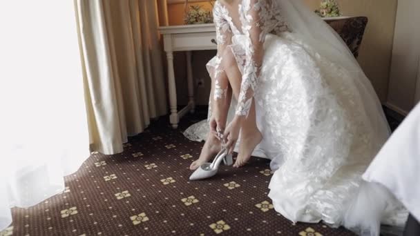 Joven novia en vestido de novia blanco con zapatos de tacón alto en piernas delgadas mientras está sentado en casa - Metraje, vídeo