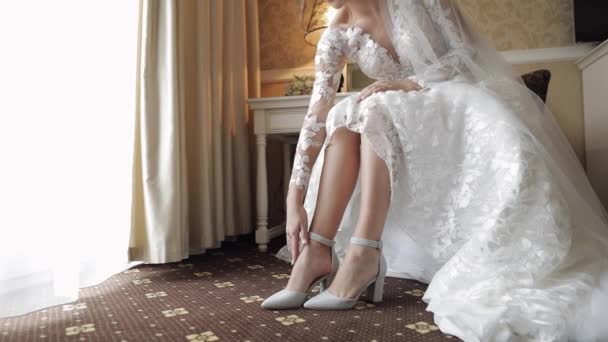 Junge Braut im weißen Hochzeitskleid trägt hochhackige Schuhe an schlanken Beinen, während sie zu Hause sitzt - Filmmaterial, Video