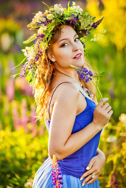 Une fille avec une couronne de fleurs sur la tête est assise au coucher du soleil dans un champ avec des fleurs de lupin violet. Image avec mise au point sélective, tonification et effet sonore - Photo, image