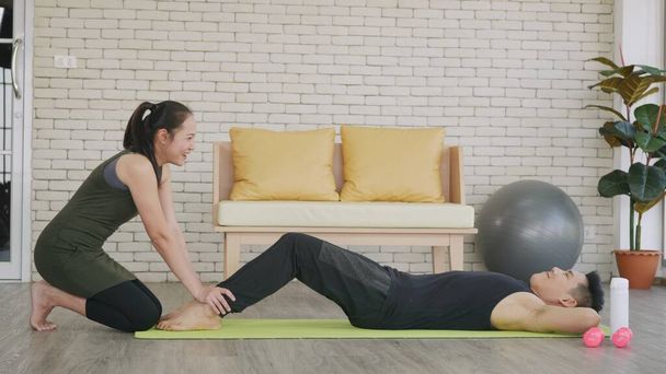 Happy Asian family couple husband and wife healthy trainer workout home in the living room. Fit man lifestyle doing sit up fitness exercise at home on floor with girlfriend support helping boyfriend - 写真・画像