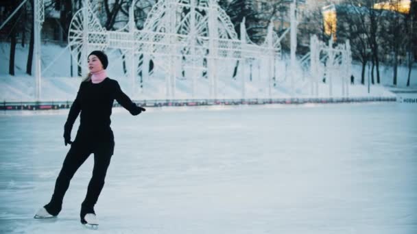 Jonge blonde vrouw in zwarte kleren met hoge paardenstaart figuur schaatsen op de openbare ijsbaan - Video