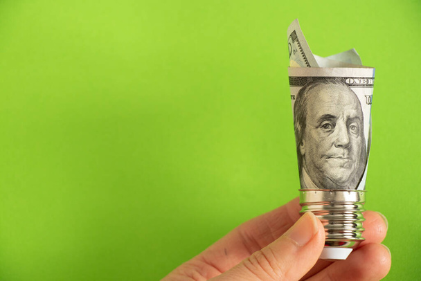 100 dolarlık banknotu olan lamba tutacağı yeşil arka plandaki bir kadının elinde vidaladı. Elektriğin fiyatı... - Fotoğraf, Görsel