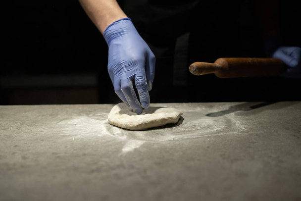 La mano de un hombre de un panadero en un guante de goma amasa masa de pizza en la mesa de trabajo. Rodillo de madera en la mano. Proceso de cocción. Lugar para su texto. - Foto, Imagen