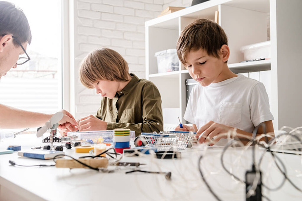 Νεαρά αγόρια και δάσκαλος διασκεδάζουν κατασκευάζοντας αυτοκίνητα ρομπότ μαζί στο εργαστήριο - Φωτογραφία, εικόνα