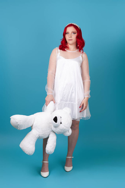 full-length sad, disappointed woman in a white dress with red hair holds a white teddy bear by the paw, isolated on a blue background - Fotografie, Obrázek