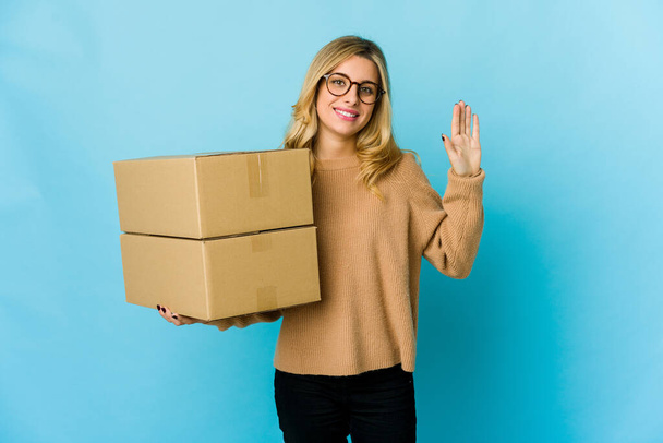 Joven rubia caucásica mujer sosteniendo cajas para mover sonriente alegre mostrando número cinco con los dedos. - Foto, imagen