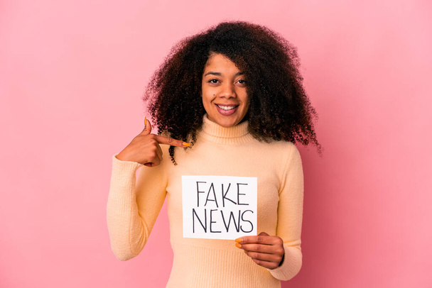 Молодая африканская кудрявая американка, держащая фальшивые новости на плакате человека, указывающего рукой на пространство для копирования рубашки, гордая и уверенная - Фото, изображение