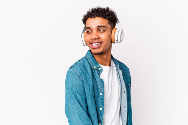 Jonge Afrikaanse amerikaanse krullende man geïsoleerd luisteren naar muziek met koptelefoon kijkt opzij glimlachend, vrolijk en aangenaam. - Foto, afbeelding