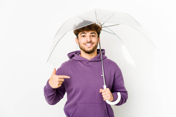 Νεαρός Άραβας κρατώντας ένα άτομο ομπρέλα δείχνοντας με το χέρι σε ένα χώρο αντιγραφής πουκάμισο, περήφανος και με αυτοπεποίθηση - Φωτογραφία, εικόνα
