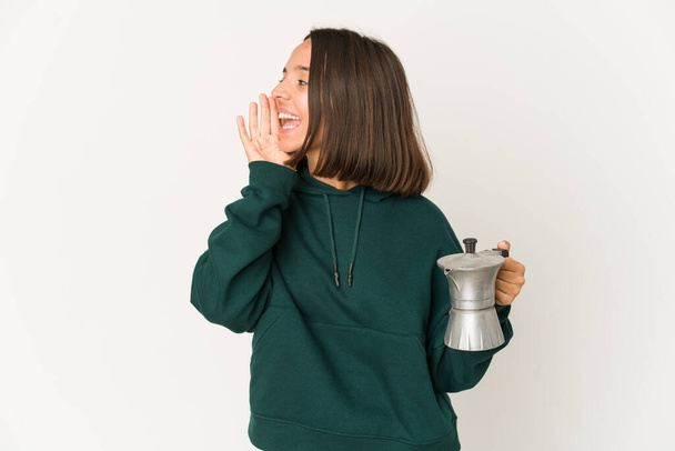 Young hispanic woman holding a coffee maker shouting and holding palm near opened mouth. - Fotoğraf, Görsel