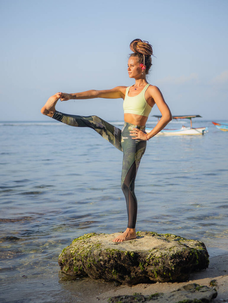 Молодая женщина практикует Utthita Hasta Padangusthasana, Standing Big Toe Hold. Тренируйся на пляже. Сильное тело. Отдых на йоге. Баланс и концентрация. Пляж Томас, Бали, Индонезия - Фото, изображение