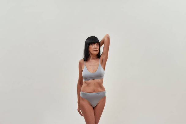 Natural beauty. Attractive half naked caucasian senior woman keeping one arm raised and looking at camera while posing in lingerie against grey background - Photo, Image