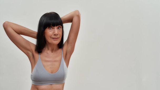 Beautiful half naked caucasian senior woman in lingerie keeping arms raised and looking at camera while standing against grey background - Foto, Imagem