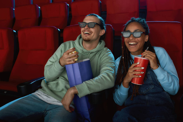 Joyful young couple wearing 3d glasses smiling, while watching a movie together in cinema auditorium, eating popcorn and drinking soda - Fotó, kép