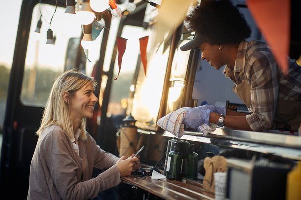 satisfied caucasian female customer talking with cheerful african employee on window of a modified truck for fast food - Фото, изображение