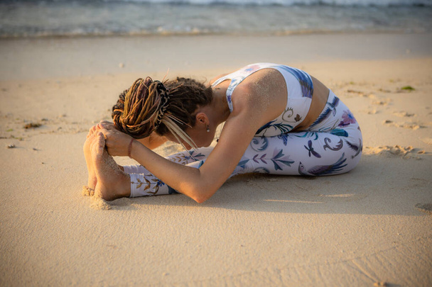 Γιόγκα παραλίας. Νεαρή λευκή γυναίκα που εξασκείται στην Πασιμοτανασάνα, καθισμένη στο μπροστινό μέρος. Ευέλικτο σώμα. Ασκήσεις τεντώματος. Υγιής σπονδυλική στήλη. Γιόγκα καταφύγιο στο Μπαλί. Αντιγραφή χώρου. Thomas beach, Ινδονησία - Φωτογραφία, εικόνα