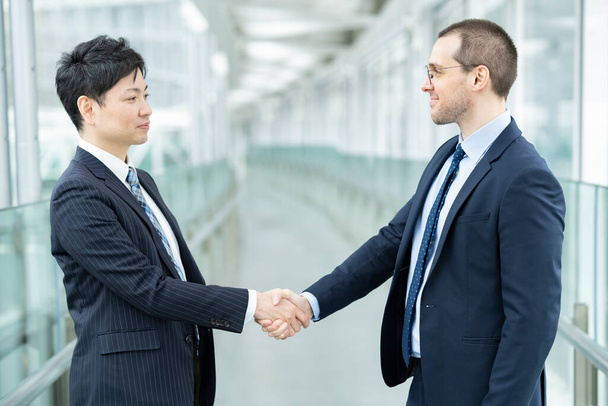 Азиатские и кавказские бизнесмены пожимают друг другу руки в костюмах - Фото, изображение