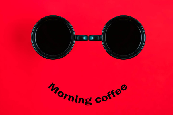 Two black coffee mugs with a smile on a red background with the phrase Morning coffee. Happy coffee mugs. - Φωτογραφία, εικόνα
