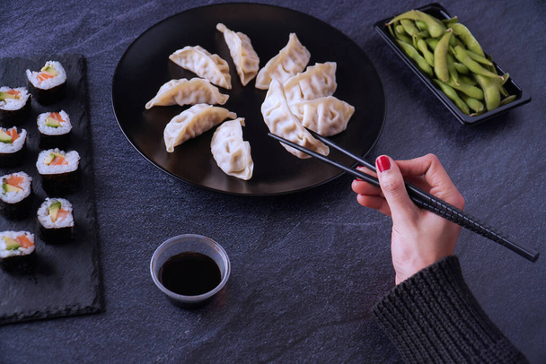 A high angle shot of a female hand about to take chicken gyozas, the salmon and avocado makis, endamame beans, and soy sauce served on the table - Valokuva, kuva