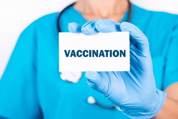 A doctor in blue medical uniform holds a card with the name of the diagnosis, the word Vaccination. Medical concept. - Valokuva, kuva