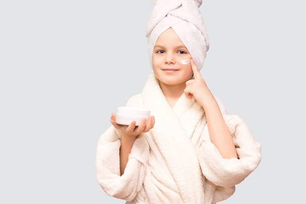 Caucasian child girl model with towel on her head puts cream on her face touching under eyes isolated on white background. - Photo, Image