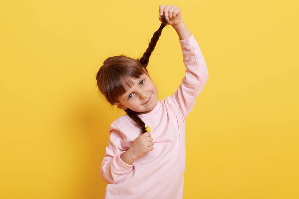 Funny little girl pulling herself in pigtails and looking directly at camera with cute smile, wearing pale pink shirt, charming child against yellow wall. - Valokuva, kuva