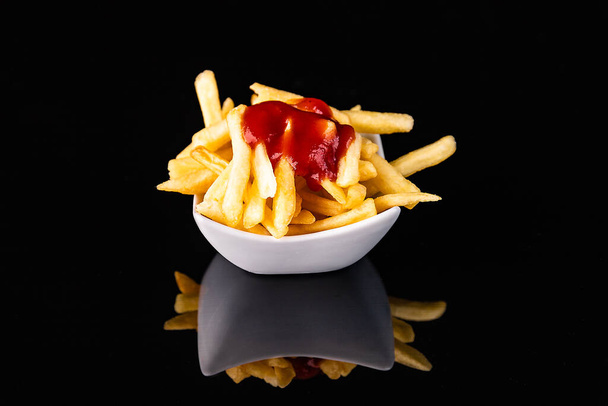 Fried potatoes piled in a plate and sprinkled with sauce, on a black background. Symbol of junk food. - Fotó, kép