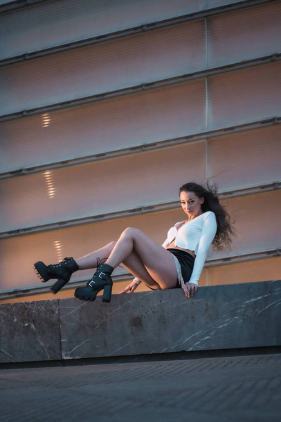 Lifestyle, a pretty brunette girl sitting in the city at the blue hour next to a gray building, wearing black boots and a short black skirt - Fotó, kép