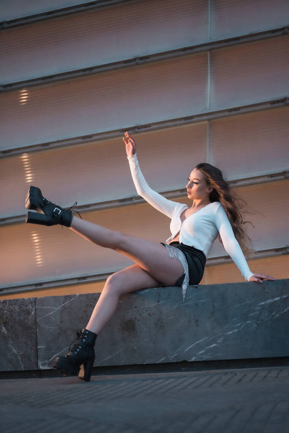 Lifestyle, posing of a pretty brunette girl sitting in the city at blue hour next to a gray building, wearing black boots and a short black skirt - Foto, imagen