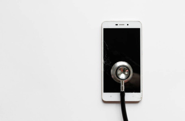 Closeup of black stethoscope on broken mobile smartphone after drop, view from above, on white background. Copy space. Smart phone with cracked screen or touchscreen and damaged case. - 写真・画像