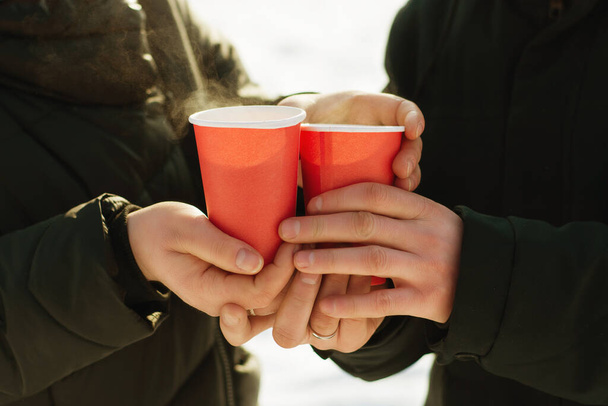 Mulled wine on the street in winter with sunny weather in the hands of a couple in love. Hot drink in disposable glasses close up - Фото, изображение