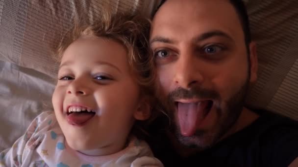 Authentieke close-up van een vader en dochter die grappige gezichten maken, kijkend naar het scherm voor een selfie, in een bed. Begrip familie en emotionele relatie tussen dochter en vader. - Video