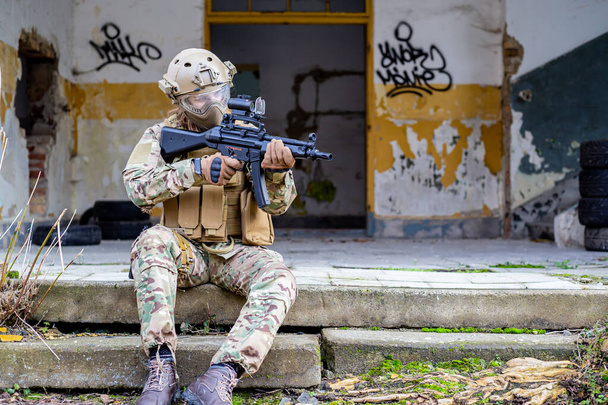 Askeri üniformalı güzel bir kız. Elinde havalı bir silahla terk edilmiş bir binanın önünde oturuyor. - Fotoğraf, Görsel