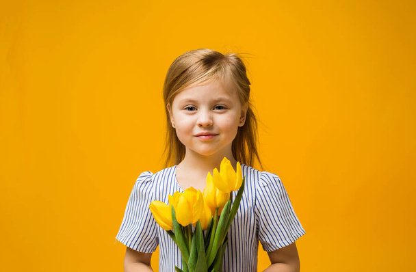 portrait of a little blonde girl with a bouquet of yellow tulips on a yellow background with space for text - Photo, Image