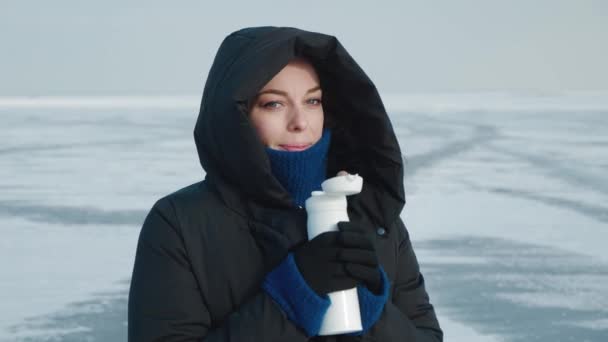Giovane donna in caldo piumino con cappuccio, beve bevanda calda da thermos in inverno freddo su sfondo artico. Ritratto di una femmina che si scalda con tè caldo sullo sfondo di lago o fiume ghiacciato - Filmati, video