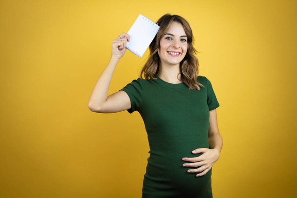 jong mooi brunette vrouw zwanger verwachten baby over geïsoleerde geel achtergrond glimlachen en tonen blanco notebook in haar hand - Foto, afbeelding
