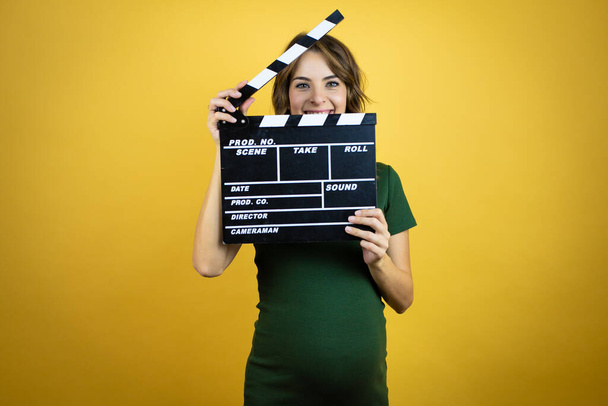 Young beautiful brunette woman pregnant expecting baby over isolated yellow background holding clapperboard very happy having fun - Zdjęcie, obraz