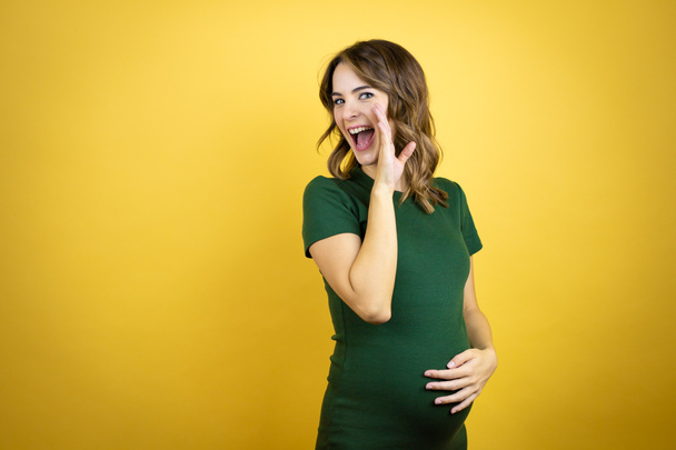 Nuori kaunis brunette nainen raskaana odottaa vauvan yli eristetty keltainen tausta käsi suulla kertoo salainen huhu, kuiskaa ilkeä puhua keskustelu - Valokuva, kuva