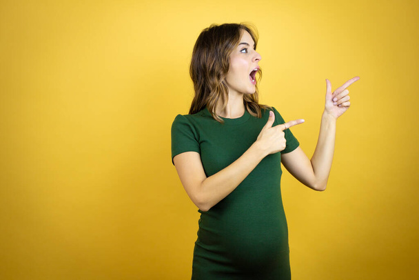 Young beautiful brunette woman pregnant expecting baby over isolated yellow background surprised and pointing her fingers side - 写真・画像