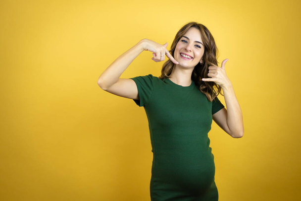 Young beautiful brunette woman pregnant expecting baby over isolated yellow background doing the call me gesture with her hands - Photo, Image