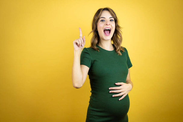 Νεαρή όμορφη μελαχρινή γυναίκα έγκυος περιμένει μωρό πάνω από απομονωμένο κίτρινο φόντο έκπληκτος και σκέφτεται με το δάχτυλό της στο κεφάλι της ότι έχει μια ιδέα. - Φωτογραφία, εικόνα
