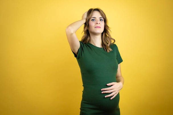 Young beautiful brunette woman pregnant expecting baby over isolated yellow background putting one hand on her head smiling like she had forgotten something - Foto, Imagem