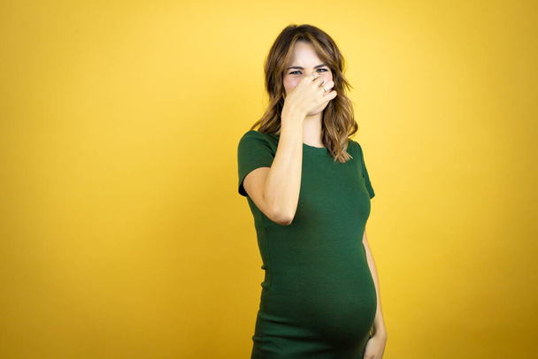 Young beautiful brunette woman pregnant expecting baby over isolated yellow background smelling something stinky and disgusting, intolerable smell, holding breath with fingers on nose - Фото, изображение