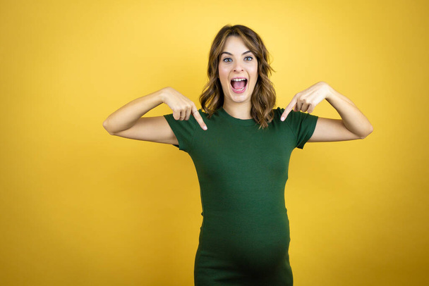 Young beautiful brunette woman pregnant expecting baby over isolated yellow background looking confident with smile on face, pointing oneself with fingers proud and happy. - 写真・画像
