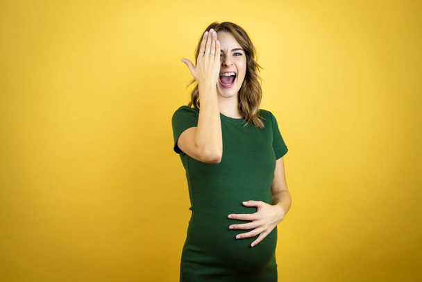 Νεαρή όμορφη μελαχρινή γυναίκα έγκυος περιμένει μωρό πάνω από απομονωμένο κίτρινο φόντο που καλύπτει το ένα μάτι με το χέρι, χαμόγελο αυτοπεποίθηση στο πρόσωπο και συγκίνηση έκπληξη. - Φωτογραφία, εικόνα
