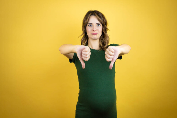Young beautiful brunette woman pregnant expecting baby over isolated yellow background with angry face, negative sign showing dislike with thumbs down - Photo, Image