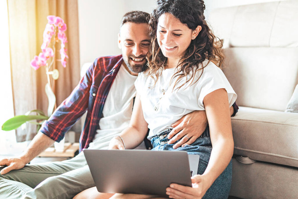 Young couple laughs and jokes while sitting on the floor browsing and shopping online with laptop - people in love search the web for new home decor - friends on video call with warm glow in background - Photo, Image